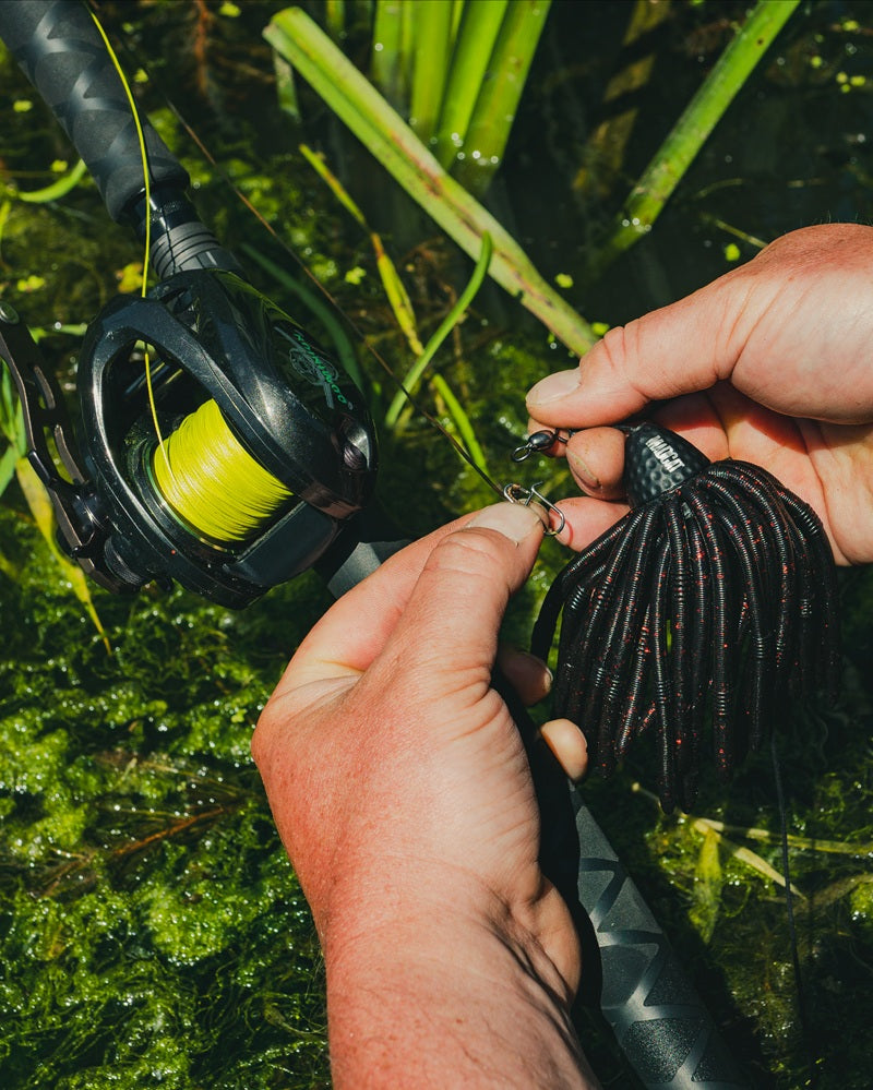 Close-up of a person's hands holding a black multi-tail fishing lure with a jig head, attached to a rod with bright yellow line, set against a background of green water plants.